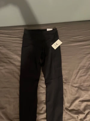 Svarta leggings från SOC, storlek S - Svarta leggings från SOC med hög midja och praktiska fickor på sidorna. Snyggt, stilrent snitt som sitter tight och följer kroppen. Perfekta för träning eller chill, tillverkade i mjukt syntetmaterial som andas. Taggen sitter kvar. Kostar (500kr) från början i butik 
