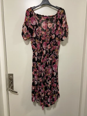 Blommig svart klänning från Indiska - Svart klänning från Indiska i fint  blommönster i på svart botten. Modellen har kort ärm, smock i midjan och vackert fall med volangkant nertill. Tunn och luftig, och passar även större byst då halsringningen är större och vid. Tyvärr hittade jag två mindre hål på kjoltyget men syns inte när man har den på sig. Se sista bild.