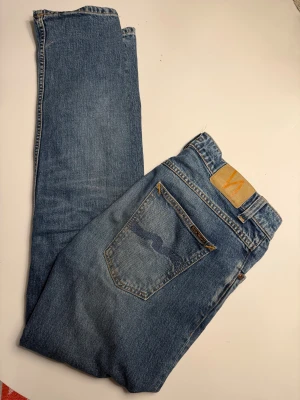 Blå jeans från Nudie Jeans - Säljer ett par klassiska blå jeans från Nudie Jeans med snygga kontrastsömmar och ikonisk broderad bakficka. Jeansen har rak passform och normal midja, tillverkade i slitstarkt bomullsmaterial. Perfekt för dig som gillar tidlös stil och kvalitetskänsla.