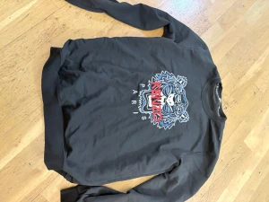 Svart Kenzo sweatshirt med tiger - Svart sweatshirt från Kenzo med ikoniskt broderat tigermotiv och texten 'KENZO PARIS' i rött och vitt framtill. Tröjan har rund halsringning och långa ärmar. Perfekt statement-plagg för dig som gillar streetstyle och snygga detaljer.