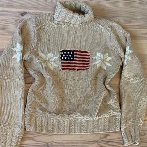 Beige stickad polotröja med flagga - Beige stickad polotröja med grov ribbad krage och muddar. På bröstet finns en amerikansk flagga i rött, vitt och blått. Tröjan har vita snöflingor på ärmar och fram, samt stickade mönsterdetaljer över hela plagget. Perfekt för kalla dagar.