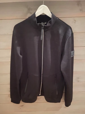 Svart zipjacka EA7 Emporio Armani - Svart zipjacka från EA7 Emporio Armani med diskret logomönster över hela tyget. Jackan har hög krage, två fickor med dragkedja och en snygg patch på ärmen. Materialet är mjukt och känns som polyester, perfekt för en chill och sportig look.