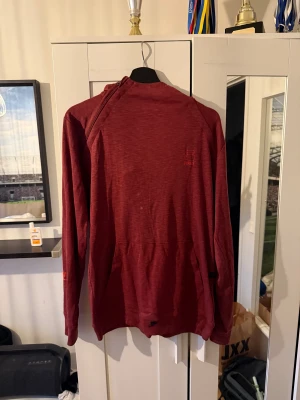 Röd hoodie från Craft med huva - Säljer en röd hoodie från Craft med lång ärm och huva. Tröjan har en enkel, sportig design med diskret logga på bröstet och ficka framtill. Perfekt för dig som gillar en avslappnad och bekväm stil.