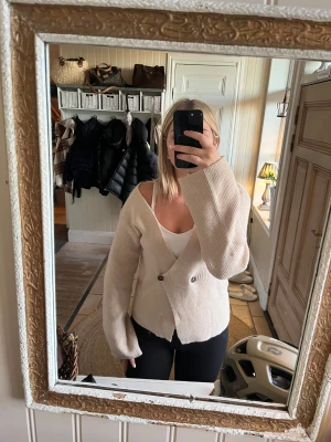 Beige stickad kofta med knappar - Säljer en beige stickad kofta med v-ringad omlottfront och två bruna knappar. Koftan har långa ärmar och en loose passform med lite längre ärmslut. Perfekt att slänga över ett linne för en chill look.