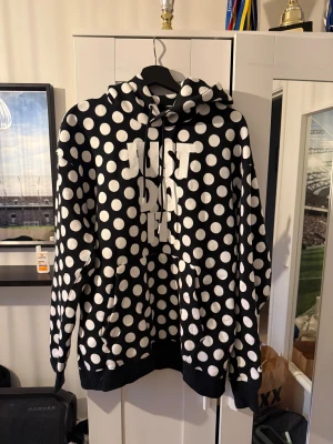 Nike hoodie med vita prickar - Svart hoodie från Nike med stora vita prickar och texten 'Just Do It' framtill. Tröjan har huva med snörning, känguruficka och långa ärmar. Perfekt för dig som gillar att sticka ut med ett lekfullt mönster och streetwear-vibe.