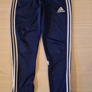 Blå Adidas träningsbyxa stl 164 - Snygga marinblå träningsbyxor från Adidas med klassiska vita ränder längs benen och logga på höften. Byxorna har dragkedjor vid fickorna och är gjorda i polyester för en sportig känsla. Perfekta för träning eller chill. Liten defekt, se bild 3. 