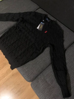 Svart kabelstickad tröja Polo Ralph Lauren - Svart kabelstickad tröja från Polo Ralph Lauren med klassisk röd broderad logga på bröstet. Tröjan har rund hals, ribbade muddar och lång ärm. Materialet är mjuk bomull som känns skönt mot huden. Perfekt för dig som gillar stilrena och tidlösa plagg. Pris kan diskuteras 