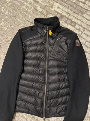 Svart pufferjacka från Parajumpers - Svart pufferjacka från Parajumpers med quiltad framsida och släta, långärmade sidor. Jackan har en gul detalj vid halsen, bröstficka med dragkedja och broderad logga på ärmen. Perfekt för dig som gillar stilren och sportig look.använd lite då och då kvitto är tyvärr borttappade men går att visa äktenhet 