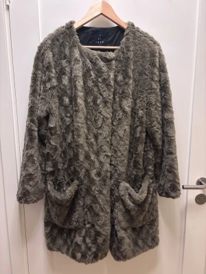 Grå pälsjacka/kappa  från Sand - Säljer en fluffig grå pälsjacka (fuskpäls) från Sand med coolt mönster och två stora fickor framtill. Jackan har lång ärm och stängs med en diskret hake. Perfekt för dig som vill ha en statement piece med skön vibe och extra mysfaktor.