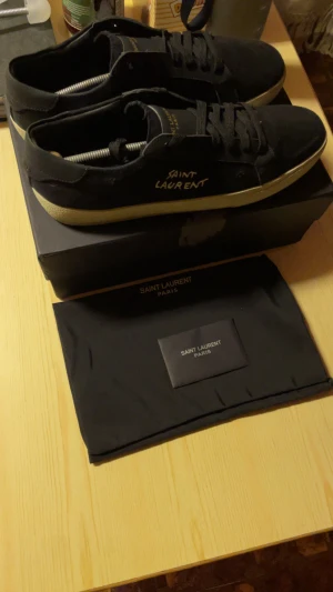 Svarta Saint Laurent sneakers - Svarta sneakers från Saint Laurent med vit logga på sidan och beige platt sula. Skorna har snörning och rund tå, tillverkade i canvas och skinn. Medföljer originalkartong och dustbag med Saint Laurent Paris-tryck. Perfekt för dig som gillar stilrena och lyxiga sneakers. 