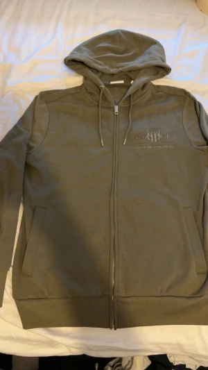 Olivgrön hoodie från GANT med dragkedja - Snygg olivgrön hoodie från GANT med broderad logga på bröstet. Tröjan har huva med snörning, två fickor fram och hel dragkedja. Mjuk insida och ribbade muddar vid ärmslut och nederkant. Perfekt för chill eller streetwear.