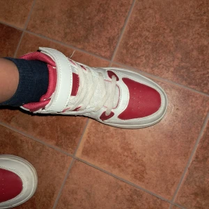 Rödvita high-top sneakers Keep Try - Säljer ett par high-top sneakers från Keep Try i rött och vitt. Skorna har snörning och kardborreband upptill för extra support. Ovandelen är i syntetmaterial med röda och vita paneler, platt sula och rund tå. Perfekt för dig som gillar sportig stil.