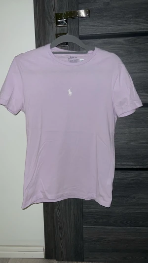 Ljusrosa t-shirt från Polo Ralph Lauren - Snygg ljusrosa t-shirt från Polo Ralph Lauren med klassisk vit logobrodering på bröstet. Modellen är custom slim fit och har rund hals samt korta ärmar. Tillverkad i mjuk bomull som känns skön mot huden. Perfekt för dig som gillar stilrena och enkla plagg.