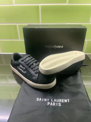 Svarta Saint Laurent sneakers - Säljer ett par svarta Saint Laurent sneakers i canvas med vita detaljer och handskriven logga på sidan. Skorna har klassisk snörning, rund tå och vit platt sula. Insidan är märkt med Saint Laurent Paris i guld. Perfekta för dig som gillar stilrena och exklusiva sneakers.