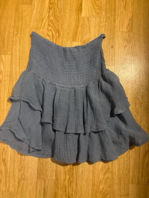 Blå volangkjol med resårmidja - Supersöt grå kortkjol med bred smock i midjan och flera lager volanger. Kjolen har en luftig och flowy känsla, perfekt för dig som gillar en avslappnad men ändå stylish look. Materialet känns mjukt och lätt mot huden.