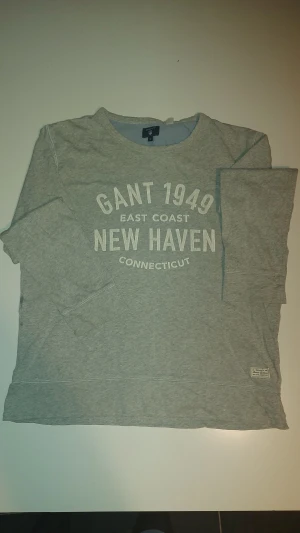 Grå långärmad tröja från GANT - Snygg grå långärmad tröja från GANT med tryck på bröstet 'GANT 1949 East Coast New Haven Connecticut'. Tröjan har rund hals och en liten patch nertill. Perfekt för dig som gillar klassisk college-stil och mjuk bomullskänsla.