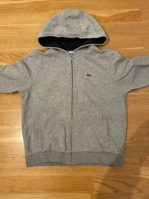 Grå hoodie från Lacoste med dragkedja - Snygg grå hoodie från Lacoste med klassisk krokodillogga på bröstet. Tröjan har huva, långa ärmar och praktisk dragkedja framtill. Tillverkad i mjuk bomullsmix och har två fickor på sidorna. Perfekt för chill dagar och streetstyle.
