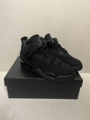Nike Air Jordan 4 Retro Black Cat - Säljer ett par Nike Air Jordan 4 Retro Black Cat i helsvart färg. Skorna har en snygg meshpanel, detaljer i mocka och skinn samt klassisk snörning. Ikonisk Jumpman-logga på plösen och Nike Air-detalj på hälen. Perfekta för dig som gillar stilrena och tidlösa sneakers.