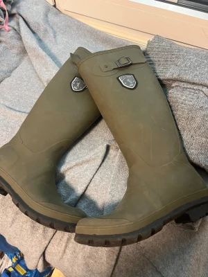 Kamik gröna gummistövlar med spänne - Säljer ett par gröna gummistövlar från Kamik med grov sula och snyggt metallspänne upptill. Stövlarna har en rund tå och ett emblem på sidan. Perfekta för regniga dagar och outdoor-äventyr. Insidan är beige och har Kamik-logga på sulan.