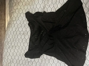 Svart blus med hål - Supersöt svart blus med volangdetaljer vid axlarna och broderade hålmönster över hela tyget. Blusen har en omlottkänsla och knytband i midjan för extra stilpoäng. Perfekt för dig som gillar en feminin och trendig look.