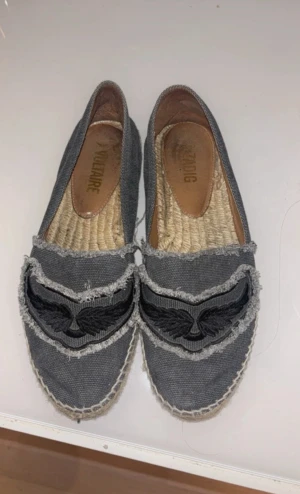Grå espadriller från Zadig & Voltaire - Säljer ett par grå espadriller från Zadig & Voltaire med fransade kanter och broderad svart vinge på ovansidan. Skorna har rund tå och platt sula i repmaterial, ovandel i grov bomullstyg och insida i naturfärgat material. Perfekt för dig som gillar coola detaljer.