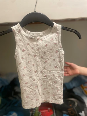 Vit blommig linnetopp från Newbie - Söt vit linnetopp från Newbie med små rosa blommor och diskret spetskant runt halsringningen. Toppen är armlös och har en mjuk bomullskänsla, perfekt för varma dagar. Blommönstret ger en gullig och fräsch vibe. Barn till tjejer, storlek 122/128