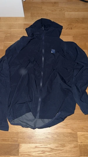 Mörkblå vindjacka från Sprayway - Snygg mörkblå vindjacka från Sprayway med huva och hel dragkedja framtill. Jackan är tillverkad i lätt syntetmaterial och har en stilren design utan mönster. Perfekt för blåsiga dagar och enkel att packa med sig. Passar dig som gillar funktion och enkel stil.