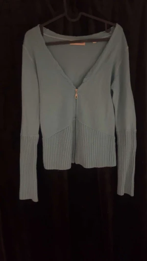 Ljusblå kofta med dragkedja Sara Kelly - Ljusblå kofta från Sara Kelly med v-ringad hals och dragkedja framtill. Tröjan har långa ärmar och breda ribbade muddar vid ärmslut och nederkant. Perfekt för dig som gillar stilrena och mjuka plagg med snygga detaljer.