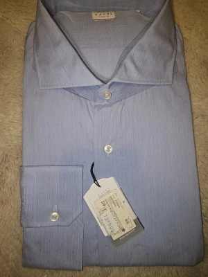 Ljusblå skjorta från Xacus - Snygg ljusblå skjorta från Xacus med diskret mönster av små X över hela tyget. Klassisk krage och vita knappar framtill samt vid manschetten. Skjortan är tillverkad i mjuk bomull och har en stilren, normal passform som funkar till många olika looks.