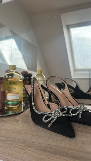 Svarta pumps med strassrosett - Snygga svarta pumps med spetsig tå och öppen häl. Dekorerade med glittrande strass runt kanten och en stor rosett framtill. Klackarna är smala och eleganta, och materialet har en satinliknande finish som ger extra lyxig känsla.