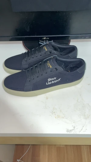 Svarta Saint Laurent sneakers - Snygga svarta sneakers från Saint Laurent med vit logga på sidan och guldfärgad text på plösen. Skorna har snörning och en beige platt sula med mönstrad undersida. Ovandelen är i textil och detaljer i skinn. Perfekt för dig som gillar stilrena och exklusiva sneakers. Kvitto finns för äkta bevis 