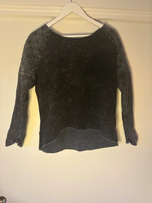 Grå stickad tröja från Tindra M - Mörkgrå stickad tröja från Tindra med quiltade axlar och små nitar som detalj. Tröjan har långärmad passform och en lite vågig nederkant för en cool look. Perfekt för dig som gillar unika detaljer och en avslappnad stil.