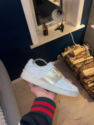 Valentino Garavani vita och guld - Säljer ett par snygga Valentino Garavani sneakers i vitt skinn med bred metallic guldrem över vristen och coola nitar på hälen. Klassisk rund tå, platt sula och snörning framtill. Perfekta för dig som vill sticka ut med lyxig streetstyle.