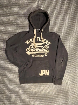 Superdry Hoodie - Svart Superdry hoodie, den ser lite blekt ut men det är bara ljuset som gör att den ser så blek ut. Defekten på sista bilden är knappast märkbar och hoodien överlag är 8/10. Om du undrar något är det bara att skriva🙌