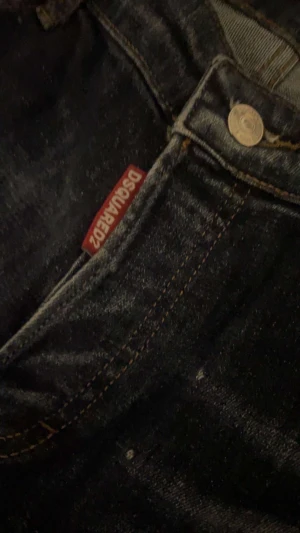 Dsquared2 blå jeans med slitningar - Säljer ett par blå jeans från Dsquared2 i storlek 54. Jeansen har coola slitningar och hål på låret, samt ljusa tvättade partier och färgstänk för en edgy look. Klassisk femficksmodell med Dsquared2-tag vid fickan och kontrastsömmar.