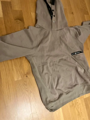 Beige hoodie från Sail Racing, M/L - Säljer en beige hoodie från Sail Racing med stor huva, knappar framtill och svart detalj vid halsen. Tröjan har en stor magficka och logga på bröstet. Perfekt för chill dagar och snygg till jeans eller joggers.