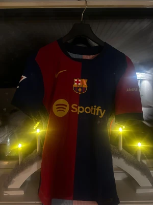 FC Barcelona Lamine Yamal matchtröja M - Säljer en officiell FC Barcelona matchtröja från Nike med Lamine Yamal och nummer 19 på ryggen. Tröjan har klassiska röda och marinblå vertikala paneler, klubbmärke på bröstet, Spotify-logga och UNHCR ACNUR-tryck. Tillverkad i lätt Dri-Fit ADV material för maximal komfort. Köptes på den officiella FC Barcelona shoppen. Nypris ca 1400-1600kr
