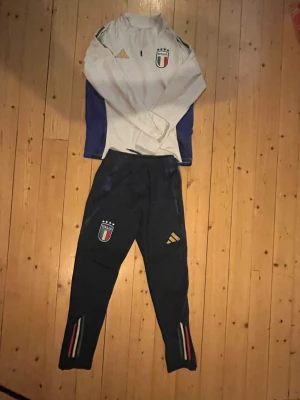 Italien Adidas half zip träningströja - Säljer en vit långärmad träningströja från Adidas med Italiens landslagsemblem på bröstet. Tröjan har half zip, gröna detaljer på axlarna och blå paneler på sidorna. Perfekt för fotbollsträning eller chill. Materialet är polyester.