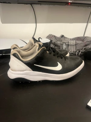Svarta Nike golfskor med vit sula - Säljer ett par svarta Nike sneakers med vit sula och vit swoosh på sidan. Skorna har svart snörning och beige meshfoder för extra komfort. Ovandelen är i syntetmaterial och designen är klassisk och sportig, perfekt för dig som gillar stilrena sneakers.