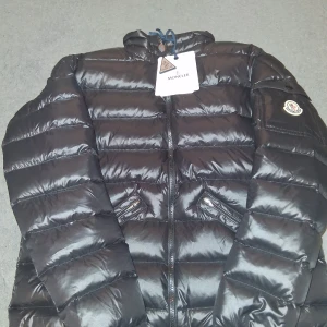 Svart Moncler pufferjacka herr - Svart pufferjacka från Moncler med glansig finish, klassisk quiltad design och dragkedja framtill. Jackan har två snedställda fickor med dragkedja och Moncler-logga på ärmen. Perfekt för kalla dagar och riktigt snygg till streetwear.