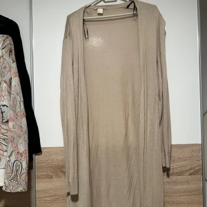Beige lång kofta från H&M - Lång och tunn beige kofta från H&M med öppen front och långa ärmar. Perfekt att slänga över en t-shirt eller linne för en chill och clean look. Materialet är mjukt och lätt, vilket gör den skön att bära under både vår och höst.