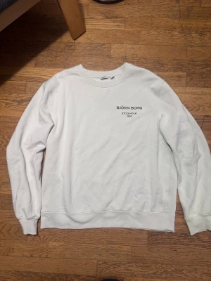 Vit sweatshirt från Björn Borg - Vit sweatshirt från Björn Borg med svart tryck på bröstet där det står 'BJÖRN BORG STOCKHOLM 1956'. Klassisk rund halsringning och långa ärmar. Mjuk insida och ribbade muddar vid ärmslut och nederkant. Perfekt för chill dagar eller när du vill ha en clean look. Färgen är vit/cremé vit.