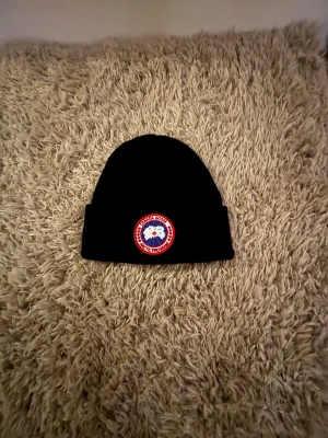 Svart mössa från Canada Goose - Svart mössa från Canada Goose med klassisk rund logotyp framtill i rött, vitt och blått. Enkel och stilren design med uppvikt kant. Perfekt för kalla dagar och snygg till streetwear. Materialet är mjukt och värmande, troligen stickad.