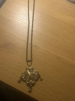 Kalevala  - Vintage Nauhasolmu riipus i brons från Kalevala Det stora Nauhasolmu-hänget designades av konstnären Germund Paaeri (1881-1950) på 1940-talet. Bandknuten inspirerades av kedjebärare dekorerade med bandfläta, som prydde kvinnors festliga dräkter i slutet av järnåldern i östra Finland och Karelen. Hängets bredd 49 mm, höjd inklusive hänglänkar 79 mm. Avigsidan är omönstrad. Ankarkedja 70 cm, inget lås.