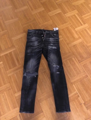 Svarta Dsquared2 jeans med slitningar - Säljer ett par svarta jeans från Dsquared2 med coola slitningar och tvättade detaljer. Modellen har knappar i gylfen och klassisk femficksdesign. Jeansen har en smal passform och är tillverkade i jeansmaterial med snyggt sliten look.