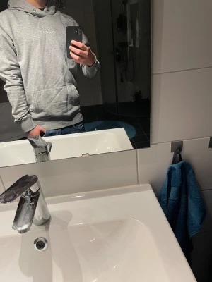 Grå hoodie från GANT med logga - Modellen är 174 och väger 61 kg. Snygg grå hoodie från GANT i storlek M. Tröjan har huva med vit kant och GANT-logga, samt en vit logga längs ena ärmen. Tillverkad i mjuk bomull och polyester, perfekt för chill dagar. Klassisk känguruficka framtill och ribbade muddar.