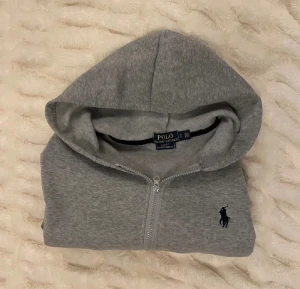 Grå zipup hoodie Polo Ralph Lauren - Snygg grå hoodie Polo Ralph Lauren med dragkedja och klassisk broderad logga på bröstet. Tröjan har huva, långa ärmar och två fickor framtill. Mjuk bomullskvalitet som är perfekt för chill dagar. Enkel och stilren design som funkar till allt. OBS pris kan diskuteras och om ni har andra frågor är det bara och ställa dem