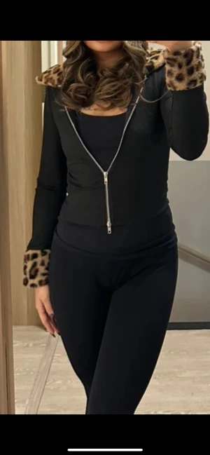 Svart cardigan jacka med leoparddetaljer - Svart cardigan jacka med dragkedja framtill och långa ärmar. Krage och ärmslut har mjuka leopardmönstrade pälsdetaljer som ger en cool och trendig vibe. Jackan är figurnära och passar perfekt till en edgy streetstyle-look.