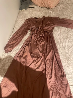 Brun långklänning med knyt från utländsk affär - Snygg långklänning i rosa nyans från en utländsk affär. Klänningen har långa puffärmar, v-ringning och ett brett knytband framtill som ger en cool detalj. Tillverkad i ett glansigt polyestertyg som faller fint. Perfekt för dig som vill sticka ut med en elegant och trendig look.