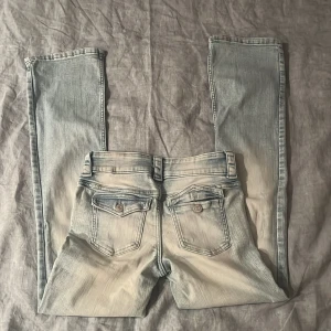 Bootcut ljusblå jeans med låg midja - Säljer ett par ljusblå bootcut jeans med låg midja och justerbar midja. Jeansen har snygga slitningar, dubbla knappar framtill och bakfickor med lock och knapp. Materialet är en mix av bomull, polyester och elastan för extra stretch.